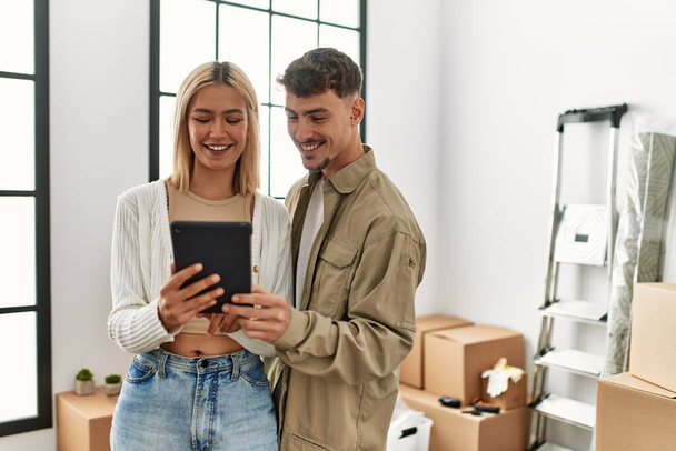 Νεαρό καυκάσιο ζευγάρι χαμογελά χαρούμενος χρησιμοποιώντας touchpad στέκεται στο νέο σπίτι. - Φωτογραφία, εικόνα