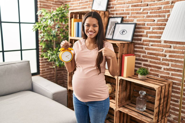 Молода вагітна жінка тримає будильник на потрібну дату, вказуючи палець на одного себе, посміхаючись щасливим і гордим
  - Фото, зображення