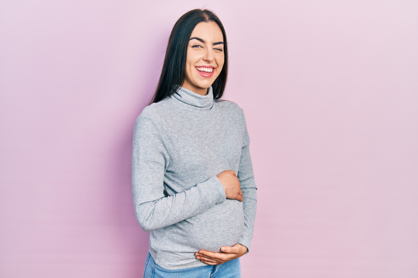 Beautiful woman with blue eyes expecting a baby, touching pregnant belly winking looking at the camera with sexy expression, cheerful and happy face.  - Zdjęcie, obraz