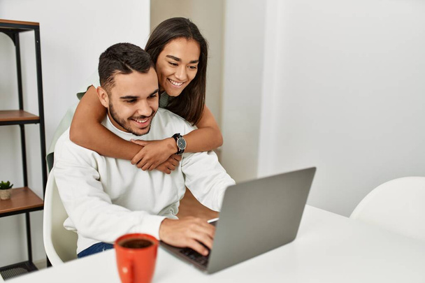 Νεαρό λατινικό ζευγάρι που χρησιμοποιεί φορητό υπολογιστή και πίνει καφέ στο σπίτι. - Φωτογραφία, εικόνα