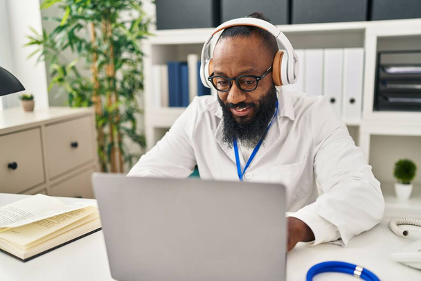 Young african american man wearing doctor uniform using laptop and headphones working at clinic - Фото, зображення
