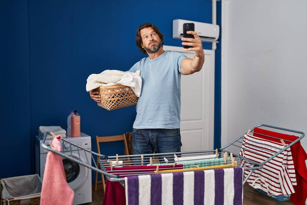Guapo hombre de mediana edad sosteniendo cesta de lavandería haciendo foto selfie hinchando mejillas con cara divertida. boca inflada con aire, captación de aire.  - Foto, Imagen