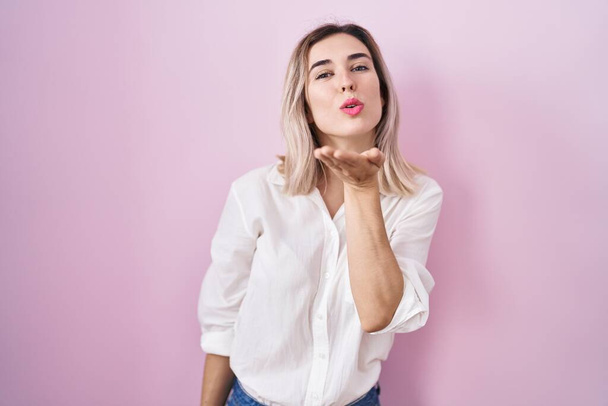 Young beautiful woman standing over pink background looking at the camera blowing a kiss with hand on air being lovely and sexy. love expression.  - Foto, Imagem