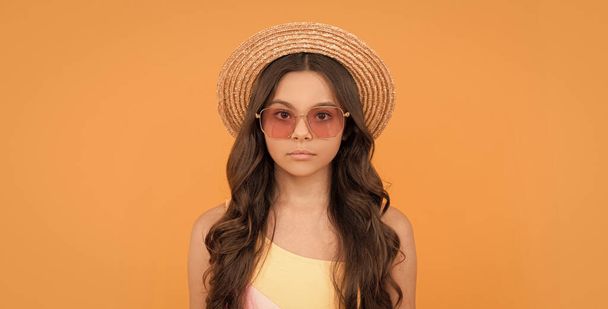 poważne dziecko w letniej słomy kapelusz i okulary ma kręcone włosy na pomarańczowym tle, modelka. - Zdjęcie, obraz