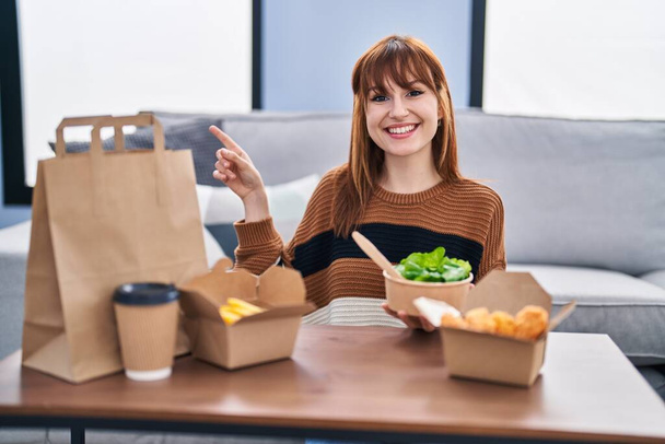 Nuori kaunis nainen syö toimitus ruokaa olohuoneessa hymyillen onnellinen osoittaa käsi ja sormi sivulle  - Valokuva, kuva