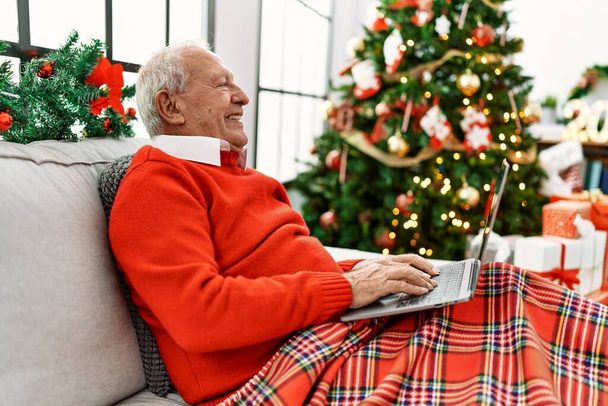 Senior uomo dai capelli grigi utilizzando il computer portatile che celebra il Natale seduto sul divano a casa. - Foto, immagini