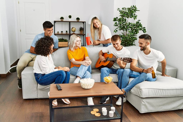 Grupo de jóvenes amigos en fiesta sonriendo feliz y tocando la guitarra clásica en casa. - Foto, imagen