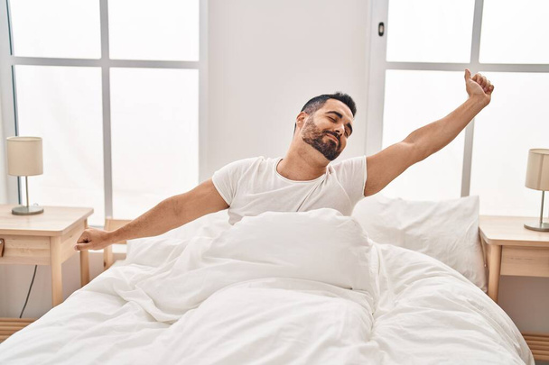 Jovem hispânico esticando os braços sentado na cama no quarto - Foto, Imagem