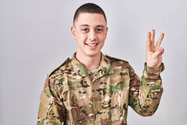 Joven vestido con camuflaje uniforme del ejército mostrando y señalando hacia arriba con los dedos número tres mientras sonríe confiado y feliz.  - Foto, Imagen