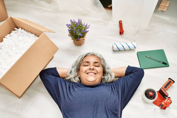 Grauhaarige Frau mittleren Alters lächelnd glücklich auf dem Boden liegend im neuen Zuhause. - Foto, Bild