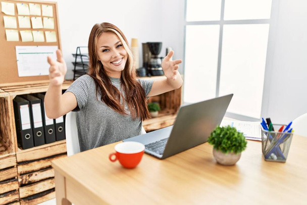 Genç esmer kadın ofiste dizüstü bilgisayarla çalışıyor kameraya sarılıp gülümsüyor. Mutluluğu kucaklayan neşeli bir ifade..  - Fotoğraf, Görsel