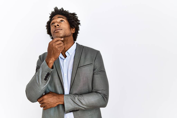 Jeune homme afro-américain vêtu d'une veste d'affaires sur fond blanc isolé visage sérieux penser à la question avec la main sur le menton, réfléchi sur l'idée déroutante  - Photo, image