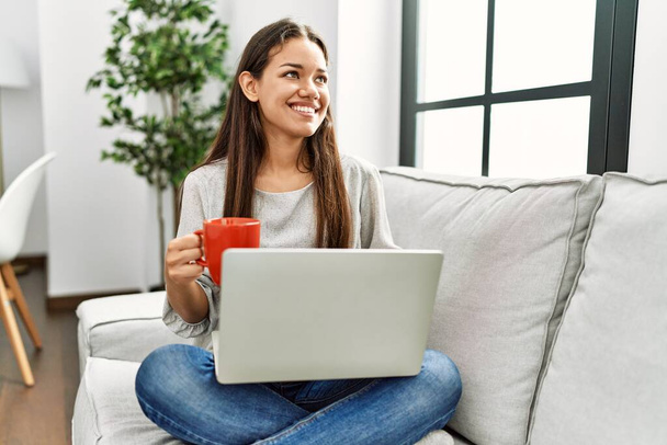 Молодая латиноамериканка с ноутбуком и пьет кофе сидя дома на диване - Фото, изображение