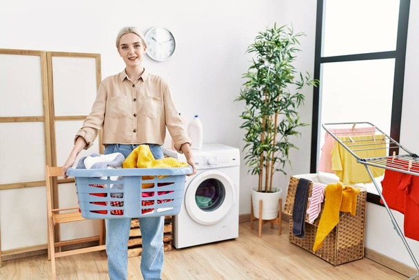 Junge Kaukasierin lächelt selbstbewusst und hält Korb mit Kleidung in der Waschküche - Foto, Bild