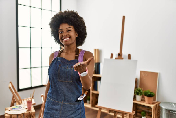 Young african american woman with afro hair at art studio smiling cheerful offering palm hand giving assistance and acceptance.  - Zdjęcie, obraz