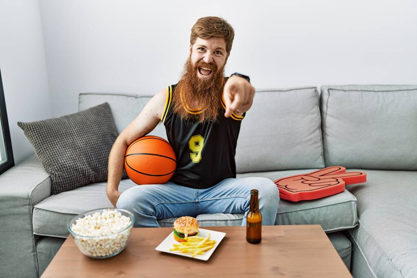 Kaukaski mężczyzna z długą brodą trzymając piłkę do koszykówki doping gra tv wskazując na Ciebie i aparat z palcami, uśmiechnięty pozytywny i wesoły  - Zdjęcie, obraz