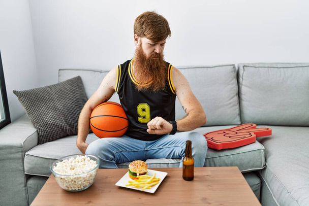 Caucasian man with long beard holding basketball ball cheering tv game checking the time on wrist watch, relaxed and confident  - Photo, image