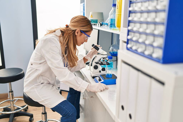 Νεαρή ξανθιά γυναίκα που φοράει επιστημονική στολή χρησιμοποιώντας μικροσκόπιο στο εργαστήριο - Φωτογραφία, εικόνα