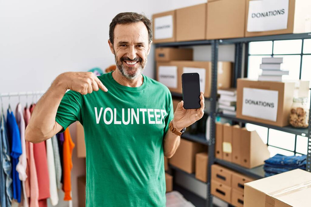 Middle age man with beard wearing volunteer t shirt holding smartphone looking confident with smile on face, pointing oneself with fingers proud and happy.  - Фото, зображення