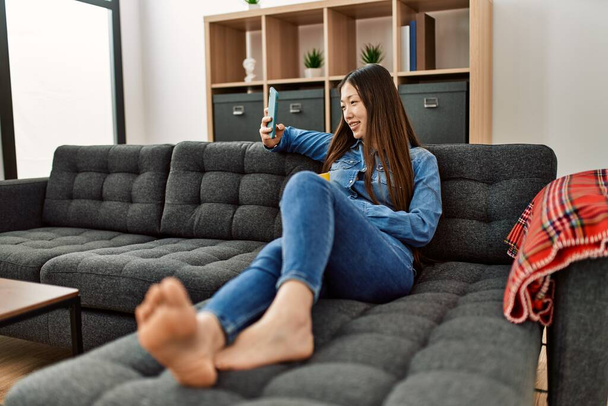 Fiatal kínai lány kávét iszik és okostelefont használ otthon a kanapén ülve.. - Fotó, kép
