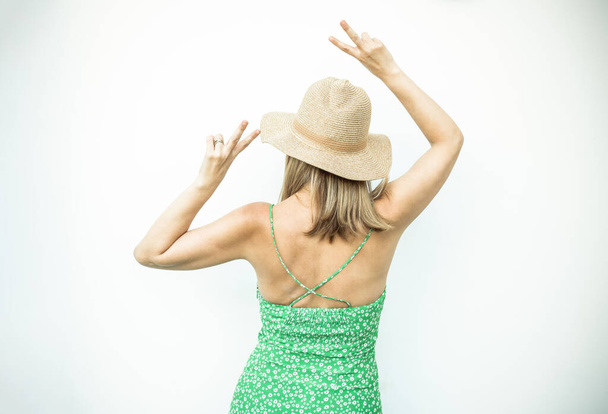 Back view young woman wearing straw hat and green dress showing peace symbol on white wall. - Foto, afbeelding