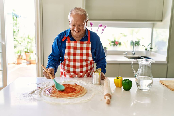 Hombre mayor sonriendo confiado cocinar pizza en la cocina - Foto, Imagen