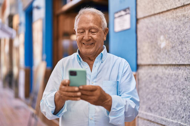 Senior siwowłosy mężczyzna uśmiecha się pewnie za pomocą smartfona na ulicy - Zdjęcie, obraz