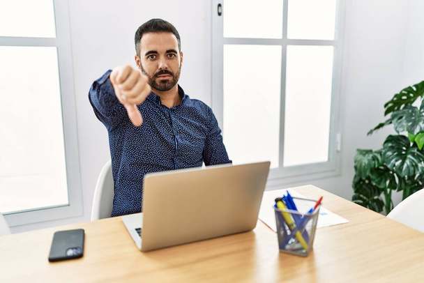 Fiatal, szakállas spanyol férfi dolgozik az irodában laptoppal boldogtalannak és dühösnek tűnik, elutasítást mutat, és negatív hüvelykujjal lefelé gesztussal. rossz kifejezés.  - Fotó, kép