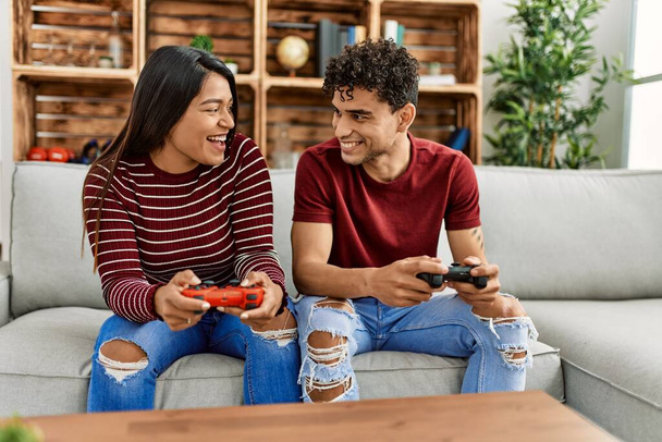 Joven pareja latina sonriendo feliz jugando video juego sentado en el sofá en casa. - Foto, Imagen