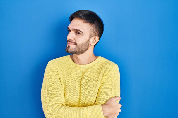 Hispanic man standing over blue background looking to the side with arms crossed convinced and confident  - Фото, изображение