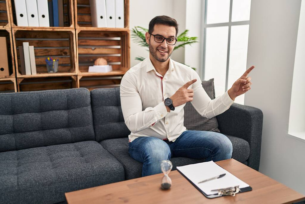Junger hispanischer Mann mit Bart, der lächelnd im Beratungsbüro arbeitet und mit zwei Händen und Fingern zur Seite in die Kamera blickt.  - Foto, Bild