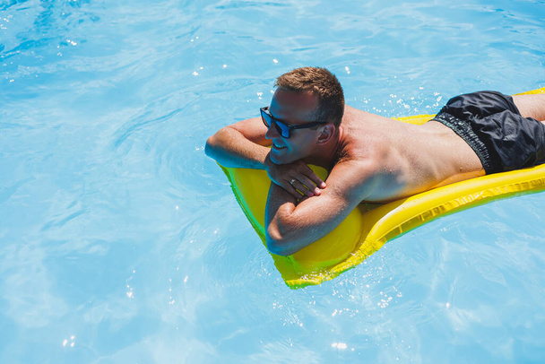 Un joven atractivo en gafas de sol y pantalones cortos se relaja en un colchón amarillo inflable en la piscina. Vacaciones de verano - Foto, imagen