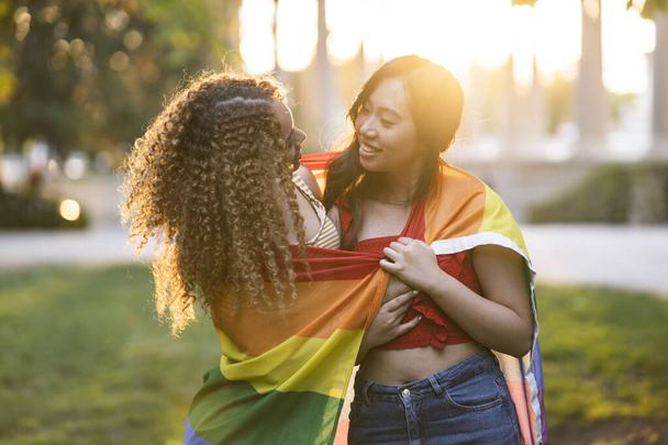 two multiethnic girlfriends with lgbt rainbow flag hugging at sunset - Foto, imagen