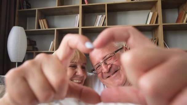 Portrait of happy elderly married couple making heart gesture with fingers showing love or showing sincere feelings together indoors, slow motion. - Footage, Video