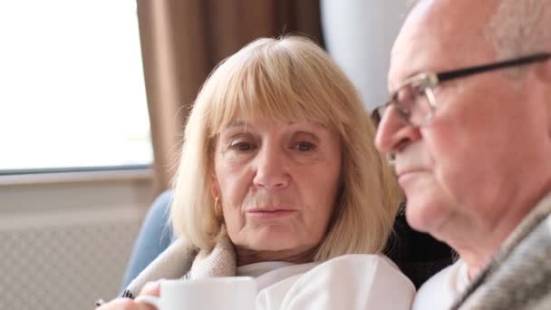Happy retired couple drinking tea and spending time together. An elderly couple is relaxing in the living room - Materiał filmowy, wideo