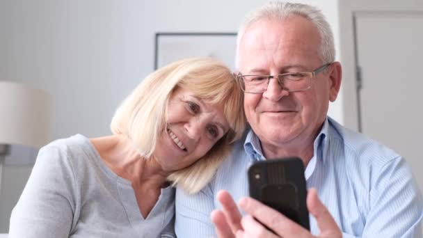 Una pareja jubilada utiliza un teléfono inteligente, sonríen mientras miran fotos divertidas en Internet. Una pareja de ancianos utiliza un aparato moderno - Metraje, vídeo