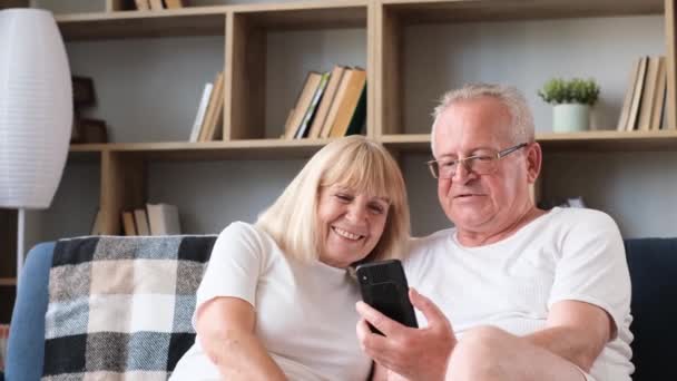 Happy family of senior couple having fun taking selfie photo together on smartphone while sitting on sofa at home. Laughing mature elderly married couple using modern gadget - 映像、動画