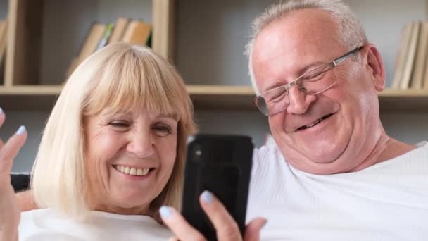 Mutlu son sınıf çifti, evdeki kanepede otururken akıllı telefondan selfie çektirirken eğleniyor. Modern aletler kullanarak yaşlı çiftlere gülüyorlar. - Video, Çekim