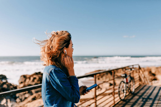 Smiling happy european woman wearing denim shirt standing on ocean shore with on ocean waves background and looking straight while listening music  - Foto, Imagen