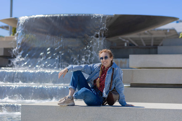 A smiling woman in a denim outfit sits in front of a cascading fountain. Rest on a warm spring day - Photo, Image