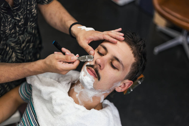 Бритва в руках специализированного парикмахера. Парикмахер бреет мужчину в парикмахерской, крупным планом. Мужские усики, бреющиеся - Фото, изображение