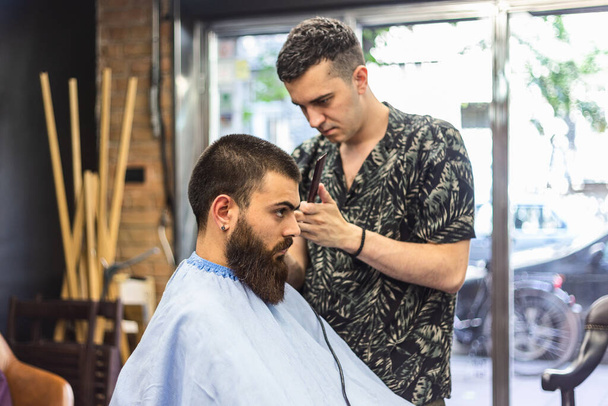 Tökéletes frizurát csinál. Fiatal szakállas férfi kap hajvágás fodrász, miközben ül székben a fodrászatban - Fotó, kép