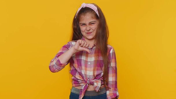 Dislike. Upset young preteen child girl kid showing thumbs down sign gesture, expressing discontent, disapproval, dissatisfied bad work indoors. Displeased toddler children on yellow studio background - Foto, Imagen