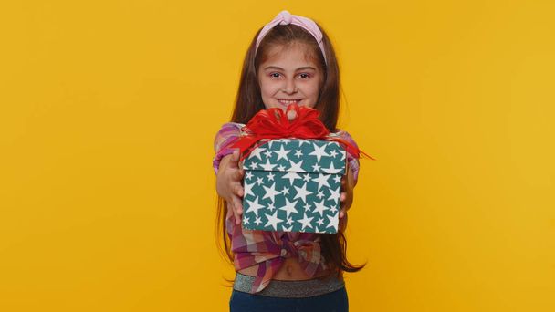 Lovely smiling young preteen child girl kid presenting birthday gift box stretches out hands, offer wrapped present career bonus, celebrating party. Little toddler children on studio yellow background - Photo, image