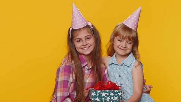 Happy little toddler children sisters girls siblings friends wearing festive cone cap celebrating birthday anniversary party, holding gift box present. Young lovely female schoolkids embracing hugging - Photo, Image