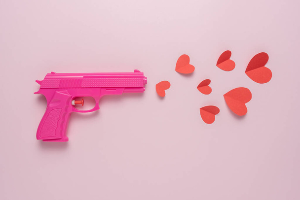 Pink handgun on pink pastel background with red paper hearts. Minimal love concept. - Fotoğraf, Görsel