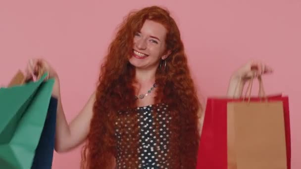 Ευτυχισμένη κοκκινομάλλα shopaholic γυναίκα δείχνει τσάντες ψώνια, διαφημιστικές εκπτώσεις, χαμογελώντας αναζητούν έκπληκτος με χαμηλές τιμές ψώνια στις διακοπές Black Friday. Τζίντζερ κορίτσι σε εσωτερικούς χώρους απομονωμένο σε ροζ φόντο - Πλάνα, βίντεο