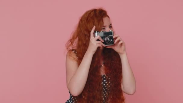 Redhead young woman tourist photographer in black dress and dress taking photos on retro camera and smiling. Travel, summer holidays vacation. Ginger girl indoors isolated on pink studio background - 映像、動画