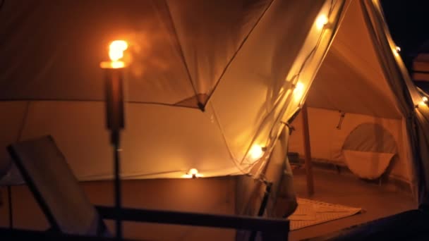 Barraca com tocha ardente, lâmpadas e cadeira de madeira em glamping, noite - Filmagem, Vídeo