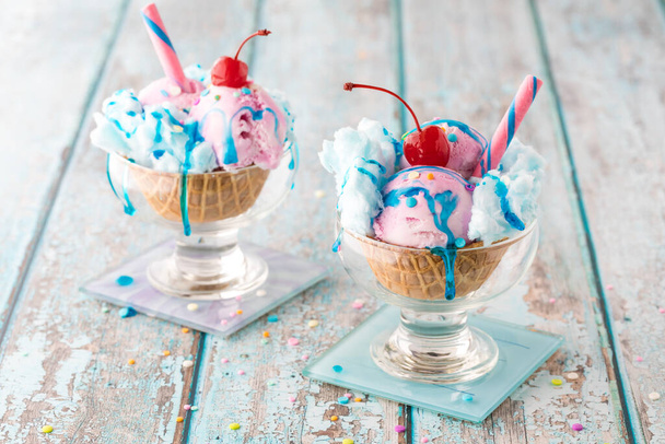 Two cotton candy sundaes in waffle bowls topped with sprinkles and syrup and garnished with a maraschino cherry and candy stick. - Zdjęcie, obraz